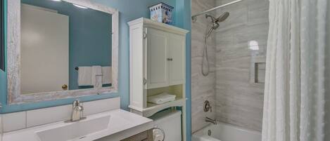 Une salle de bain avec un lavabo blanc, un miroir, des toilettes, une douche avec un rideau et un meuble de rangement.