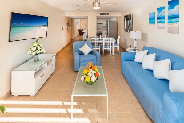 A living room with a blue sofa, a coffee table with a floral arrangement, and a dining area with a white table and chairs.