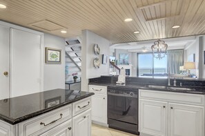 Une cuisine moderne avec des meubles blancs, un plan de travail noir et un évier en acier inoxydable. Un escalier mène à un balcon offrant une vue sur l’océan.