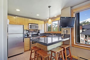 A modern kitchen with wooden cabinets, stainless steel appliances, a large island with stools, and a TV mounted on the wall. - Downtown Studio w/ Sunny Alpine Views- Sleeps 4! (Breckenridge)