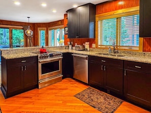 A modern kitchen with dark wood cabinets, granite countertops, stainless steel appliances, and large windows with blinds.