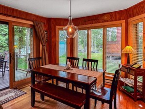 A dining area with a wooden table and chairs, a hanging light fixture, and a view through large windows.
