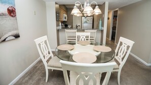A dining area with a glass table surrounded by white chairs, overhead pendant lights, and a kitchen in the background.