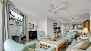 Un salon spacieux doté d’un grand téléviseur à écran plat fixé au mur, d’un ventilateur de plafond et offrant une vue sur la cuisine grâce à une porte ouverte.