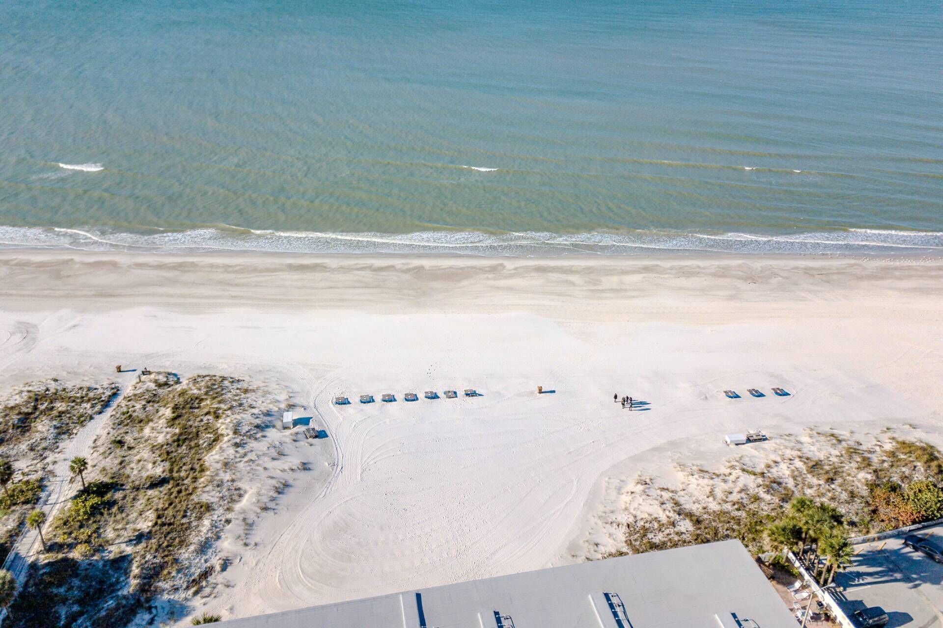 An aerial view of a beach with rows of blue beach umbrellas and lounge chairs, a parking area, and a building with a flat roof.