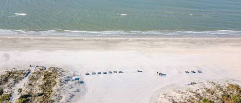 An aerial view of a beach with rows of blue beach umbrellas and lounge chairs, a parking area, and a building with a flat roof.