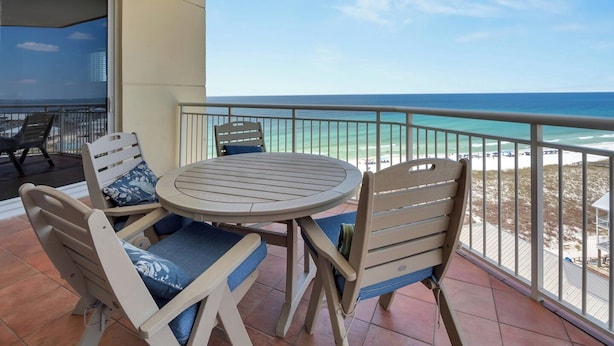 A balcony with a round table and chairs, overlooking a beach with clear blue water and a distant shoreline. - Coastal Castle. 4 Bedroom Gulf Front condo. (Navarre Beach)