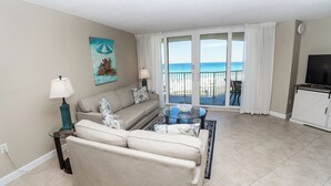 A living room with a sofa, a glass coffee table, and a TV. There is a balcony with a view of the ocean. - E604 Amazing Gulf View, 3 Pools, 2 FREE Beach Chairs! (Panama City Beach)