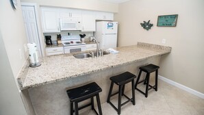 A kitchen with white cabinets, a granite countertop, a sink, a stove, and three black stools. - E604 Amazing Gulf View, 3 Pools, 2 FREE Beach Chairs! (Panama City Beach)
