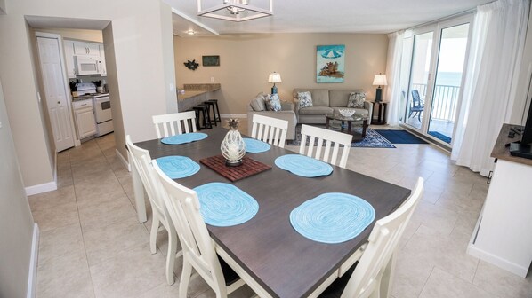 A dining area with a dark wooden table, white chairs, and blue placemats. There is a kitchen in the background with white cabinets and a microwave.
