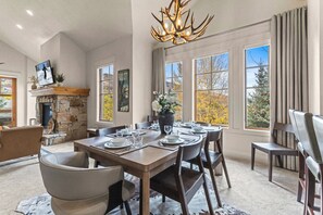 A dining area with a stone fireplace, a chandelier, and a view of autumn foliage through large windows.