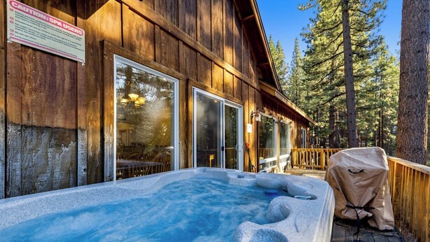 A wooden cabin with a hot tub, large windows, and a deck overlooking a forest. - Built for Fun! Spacious Mountain Home with Massive Game Room, Pool Table!- 1800HM~ (South Lake Tahoe)