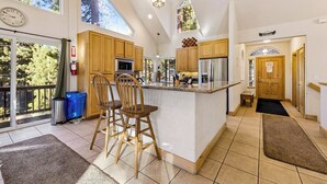 A spacious kitchen with wooden cabinets, a large island with bar stools, a clock, and a view of trees through the windows. - Built for Fun! Spacious Mountain Home with Massive Game Room, Pool Table!- 1800HM~ (South Lake Tahoe)