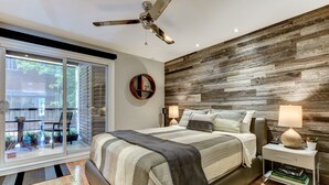 A modern bedroom with a large bed, a ceiling fan, a wooden accent wall, a round mirror, and a bedside table with a lamp. - Modern Montreal Village Condo – Chic Design, Prime Location, Incredible Style! (Centre-Sud South)