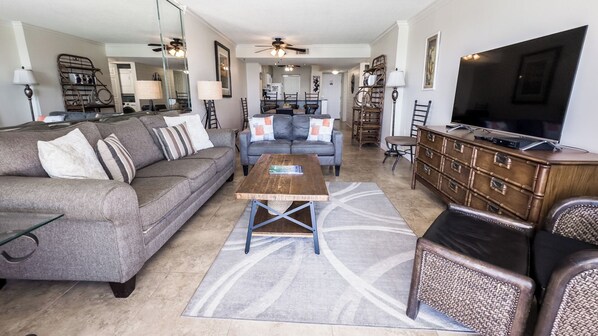 A spacious living room with a sofa, coffee table, and a television. There is a kitchen area in the background.
