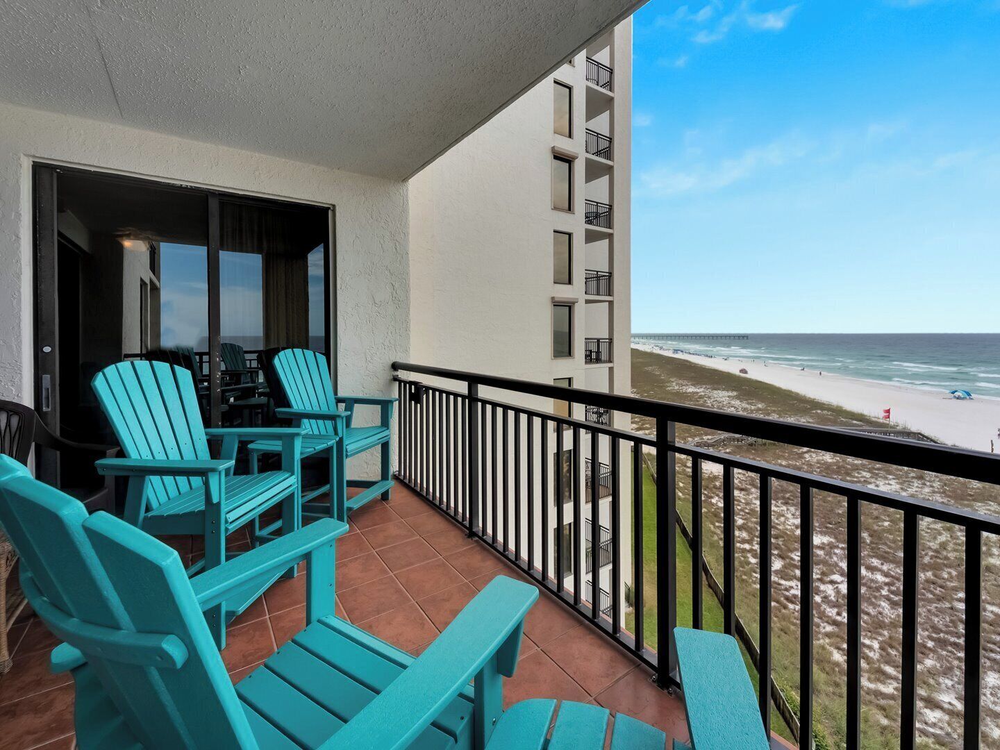 A balcony with turquoise chairs and a table, overlooking a beach and ocean.