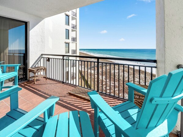 Un balcon avec des chaises turquoise, une table en osier et une vue sur la plage et l’océan.