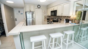 Una cocina moderna con armarios blancos, un refrigerador de acero inoxidable, un microondas y un lavamanos. Hay tres taburetes blancos y un reloj en la pared.