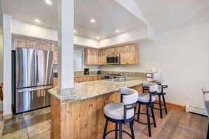 A modern kitchen with a stainless steel refrigerator, wooden cabinets, a granite countertop, and bar stools.