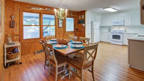 A wooden dining table with chairs, a chandelier, and a kitchen with white cabinets and appliances. - 12 42nd Avenue by AvantStay | 2 Mins To The Beach | Screened In Porch! (Isle of Palms)