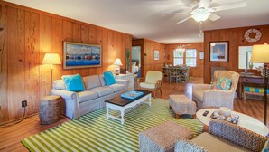 A cozy living room with wooden walls, a ceiling fan, a dining area, and a fireplace. - 12 42nd Avenue by AvantStay | 2 Mins To The Beach | Screened In Porch! (Isle of Palms)