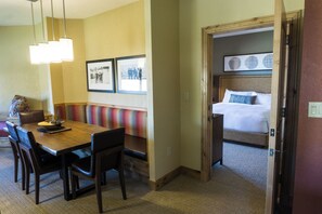 A hotel room with a wooden dining table, chairs, a bench, a bed with a white mattress and blue pillows, and a wall with framed pictures.