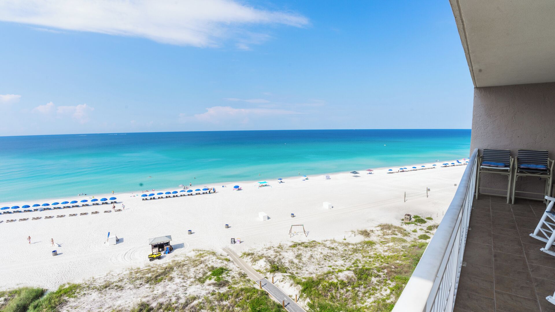 A beach view from a balcony with a clear sky, a sandy beach, and a few umbrellas.