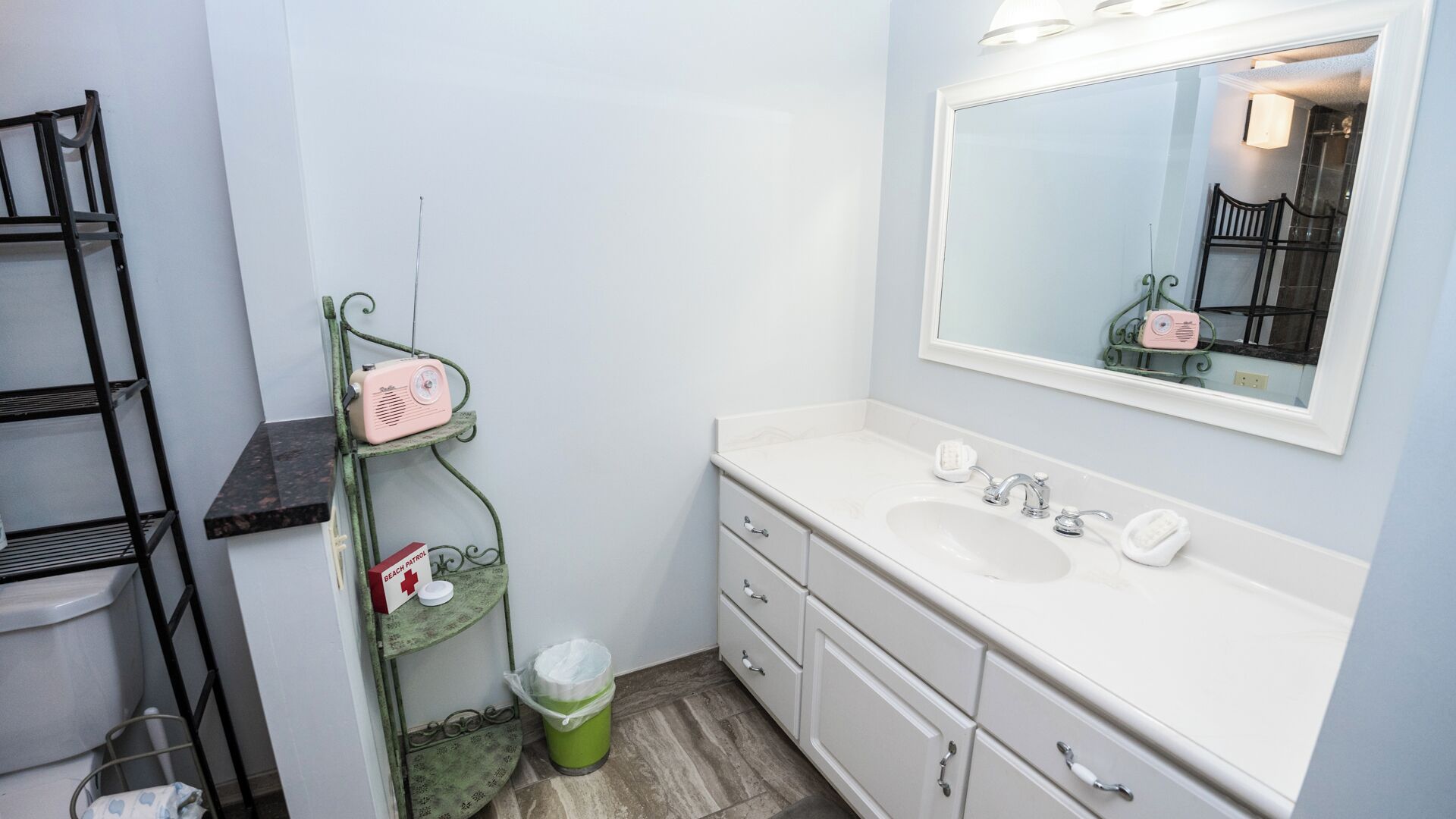 A bathroom with a white sink, a mirror, a green metal shelf with a radio, and a trash can.