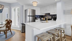 Una cocina moderna con armarios blancos, un refrigerador de acero inoxidable, una isla central con taburetes y una lámpara de araña.