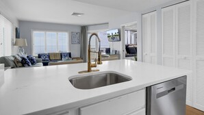 Una cocina moderna con encimera blanca, pileta de acero inoxidable y grifo dorado. Hay una zona de comedor con un sofá y un televisor al fondo.