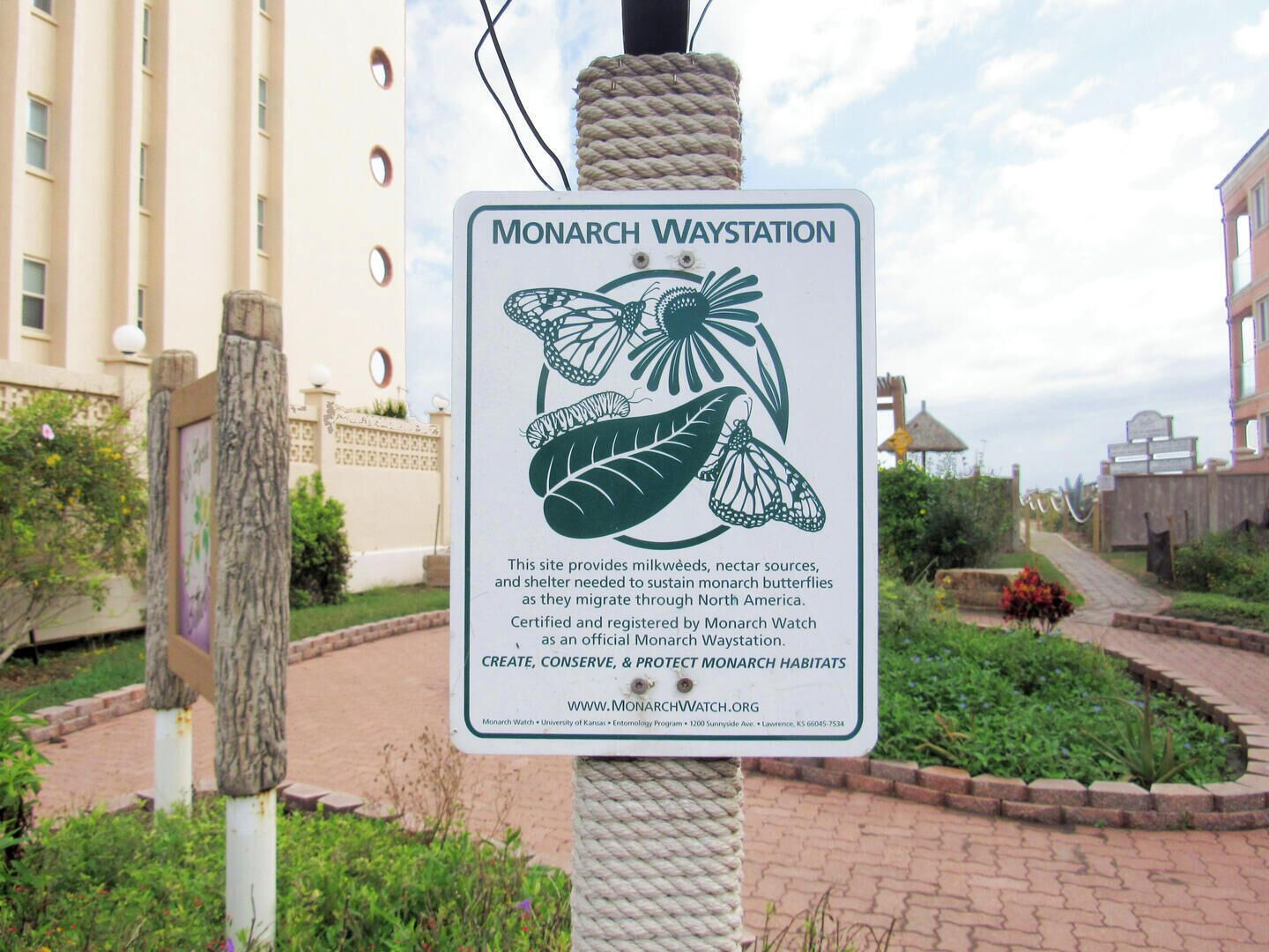 Schild für eine Monarch-Station mit Schmetterlingsabbildung, in einem begrünten Bereich mit einem Gebäude im Hintergrund.