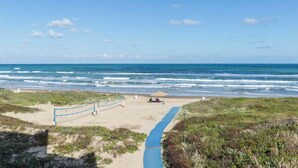 Una playa con un camino azul que lleva al agua, una zona arenosa con una cerca azul y una vista despejada del océano.