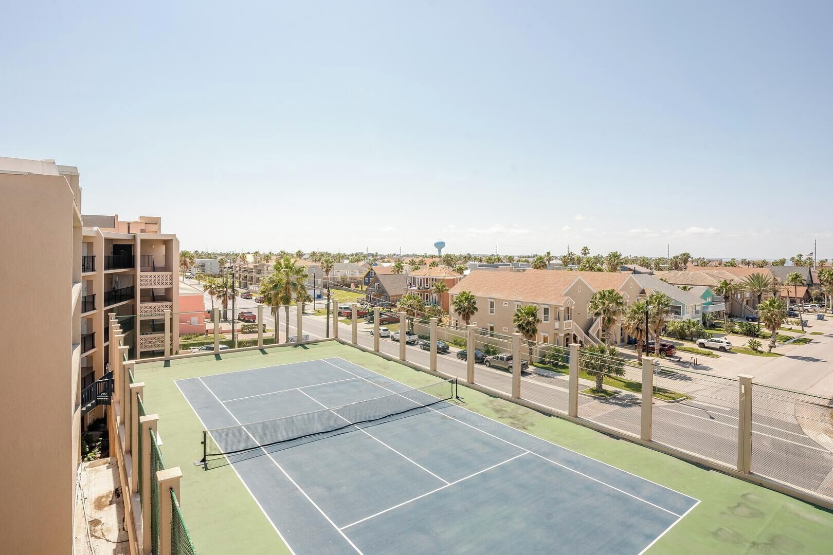 A tennis court with a net, surrounded by a fence, and buildings in the background.