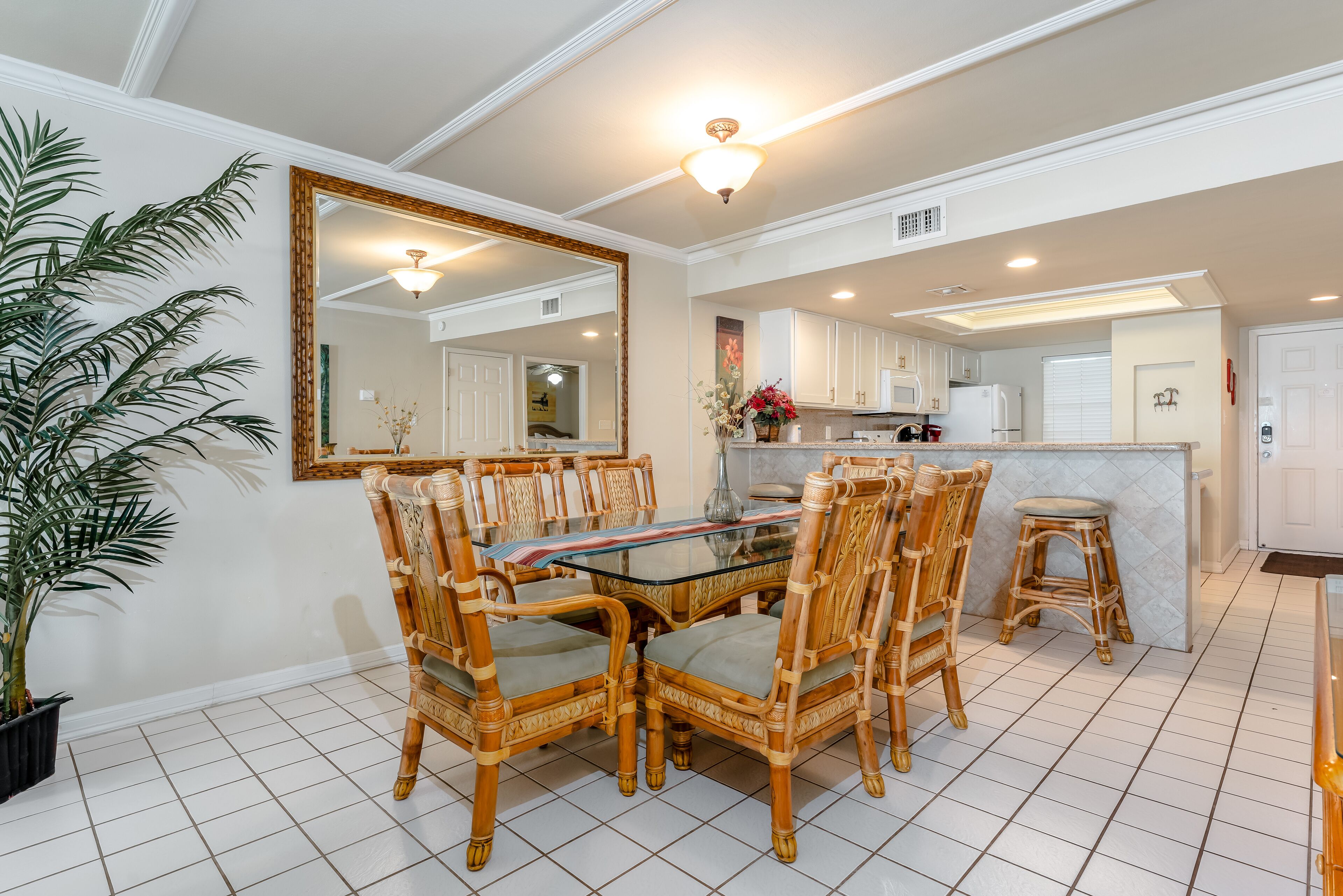A dining area with a glass table and wicker chairs, a large mirror, and a potted plant.