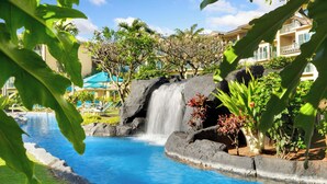 A pool with a waterfall, surrounded by tropical plants and a building with balconies. - Waipouli Beach Resort Exquisite Luxury Garden View Large Yard Perfect for Fam (Kapaa)