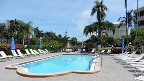 A swimming pool with lounge chairs and umbrellas, surrounded by palm trees and a building in the background.
