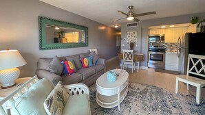 A living room with a sofa, a coffee table, a lamp, a ceiling fan, and a kitchen area with cabinets and appliances.