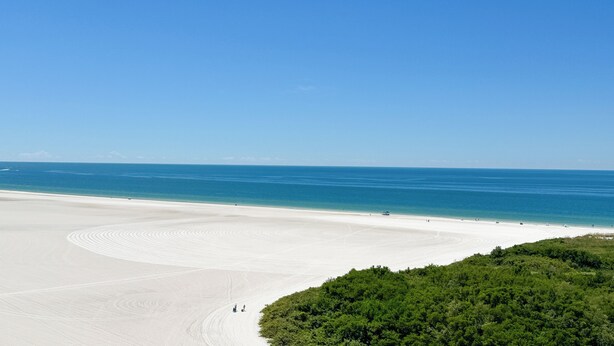 A wide sandy beach with a clear blue ocean and a forested area in the foreground. - South Seas T3-1509: Beautifully Updated Condo;  What a View! (Marco Island)