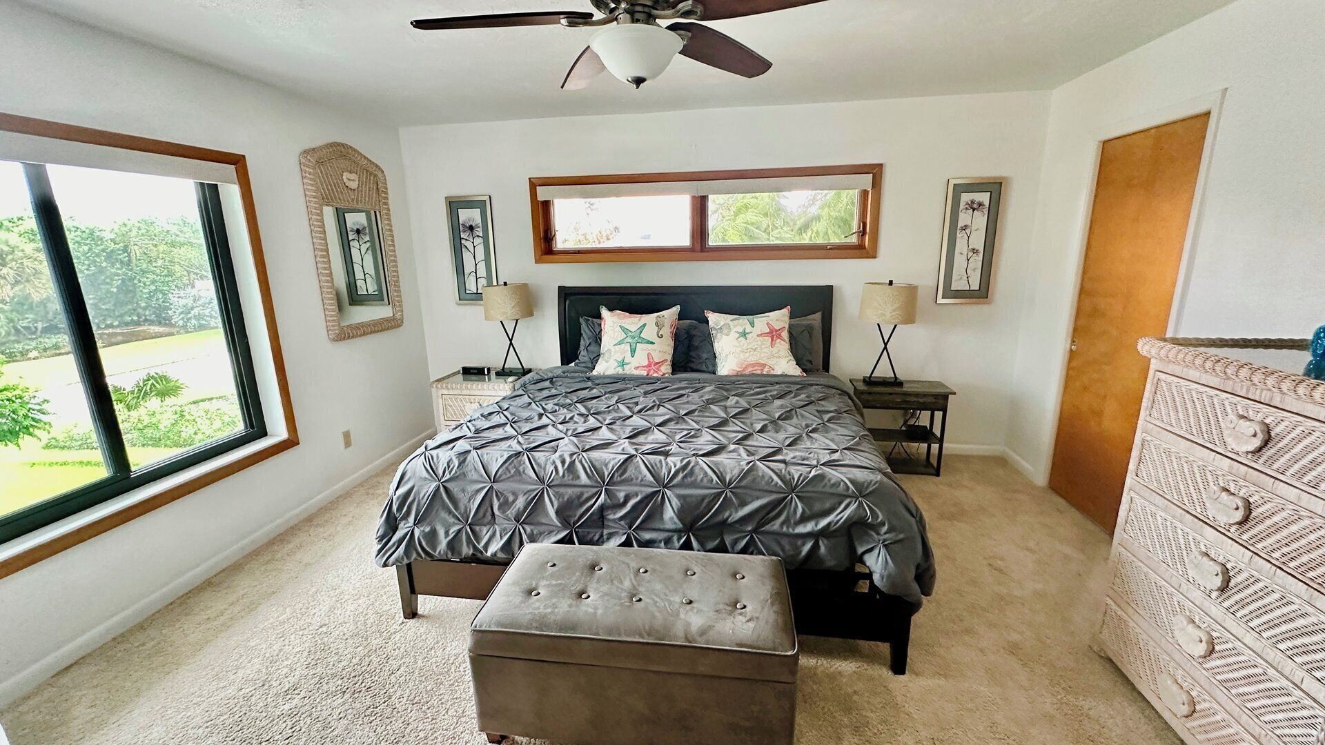 A bedroom with a large bed, a ceiling fan, a dresser, and a view of the outdoors.