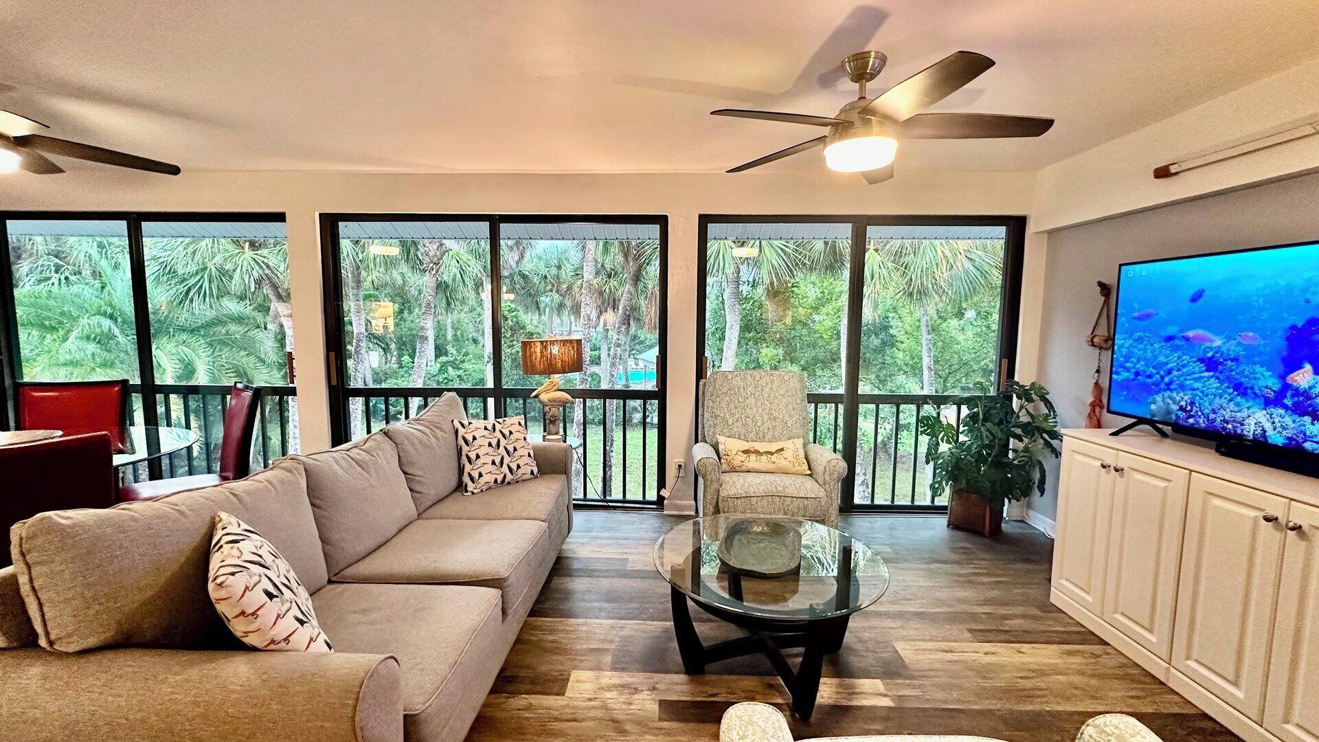 A living room with a sofa, armchair, glass coffee table, and a TV on a cabinet. Large windows offer a view of greenery outside.