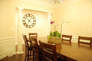 A dining room with a round table, wooden chairs, a clock, and a vase with flowers. - Spacious 5BR House, 2 pools & pickle ball, bike to beach, linen provided (Rehoboth Beach)