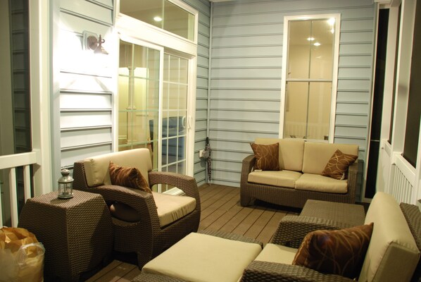A cozy outdoor seating area with wicker furniture and cushioned chairs, a small table with a lantern, and a glass door leading to a covered porch. - Spacious 5BR House, 2 pools & pickle ball, bike to beach, linen provided (Rehoboth Beach)