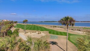 A golf course with a water body in the background, palm trees, and a clear sky. - 73 Grand Pavilion by AvantStay | Corner Lot, Porch (Isle Of Palms)