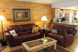 A cozy living room with two leather sofas, a glass coffee table, and a wooden wall with a framed picture.