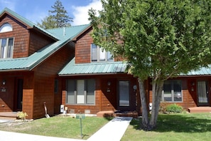 A two-story wooden building with a green roof, numbered units, and a tree in the front yard.