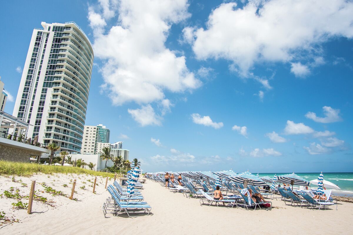 A beach with rows of sunbeds and umbrellas, a tall hotel, and a clear blue sky.