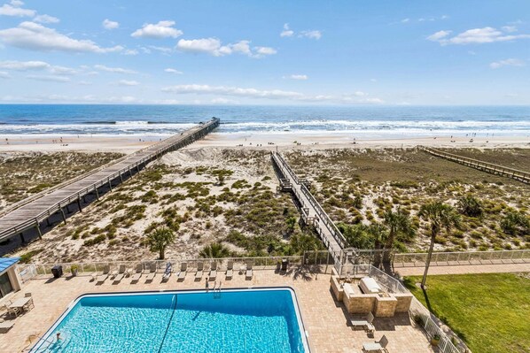 A beach view with a long pier, a swimming pool, and lounge chairs.