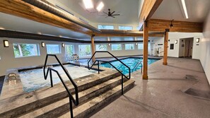 En indendørs swimmingpool med trin ned i den, træbjælker i loftet og vinduer med udsigt til træer.