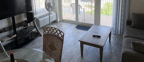 A living room with a flat-screen TV, a standing fan, a wicker chair, a wooden table, and a sofa.