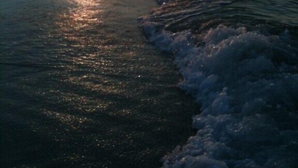 Una spiaggia al tramonto, con il sole che scompare sull'oceano, le onde che lambiscono dolcemente la riva e un molo che si protende verso l'acqua.